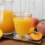 A glass of peach juice with a peach slice on the rim, sunlight streaming through the glass.