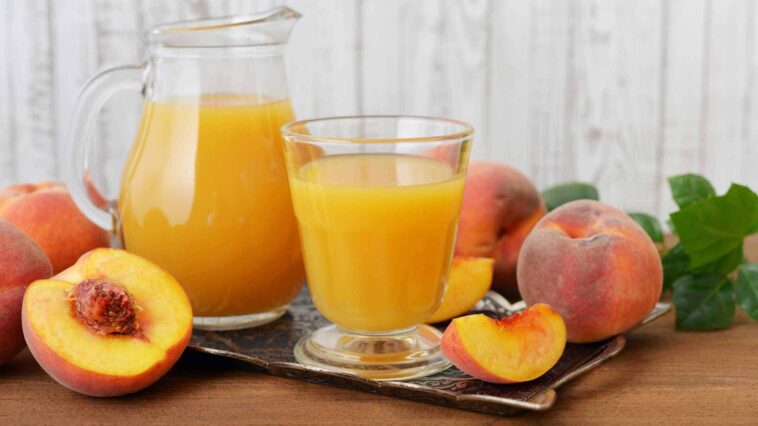 A glass of peach juice with a peach slice on the rim, sunlight streaming through the glass.